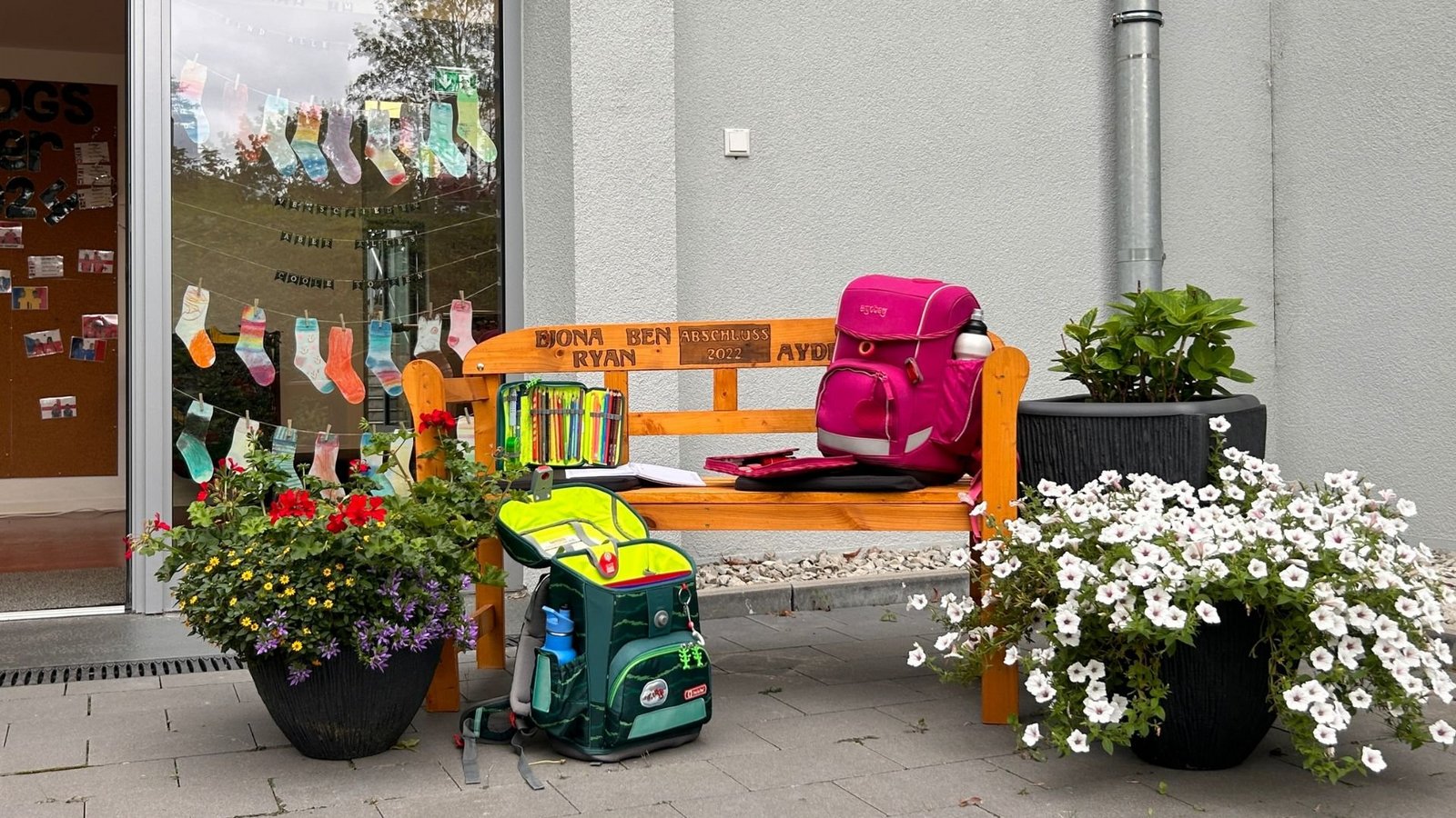 Eine Holzbank vor einer Schule mit zwei Schulranzen und bunten Blumen. Im Hintergrund eine Pinnwand mit Fotos und die Aufschrift