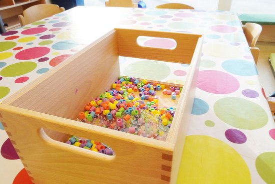 Bastelmaterial in einer Holzkiste auf einem Tisch mit bunter Tischdecke in der Kita Bunte Welt
