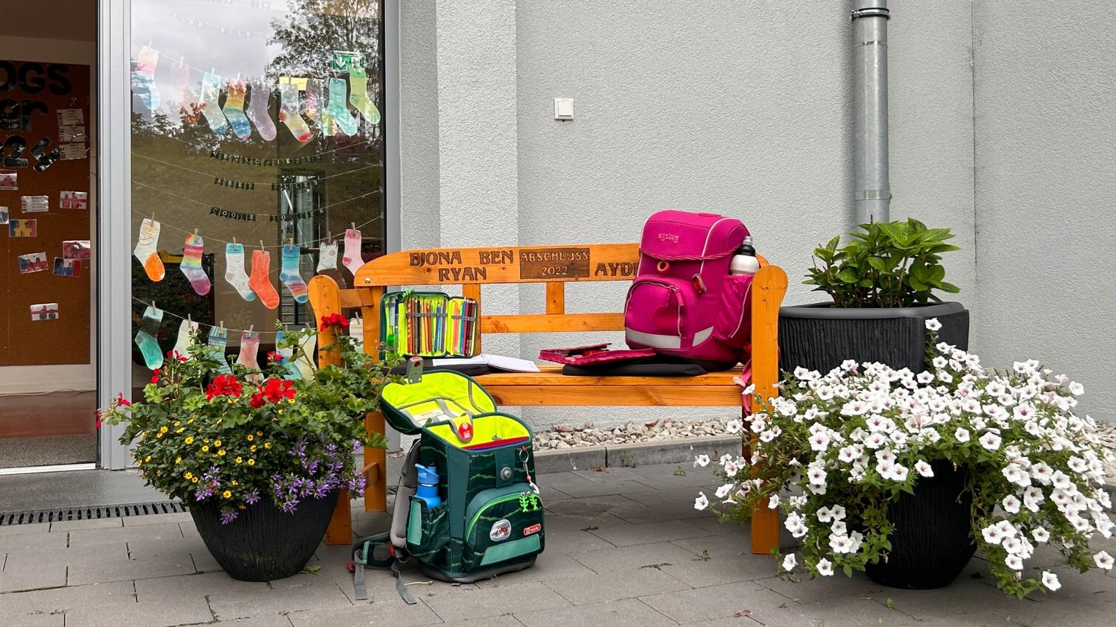 Eine Holzbank vor einer Schule mit zwei Schulranzen und bunten Blumen. Im Hintergrund eine Pinnwand mit Fotos und die Aufschrift